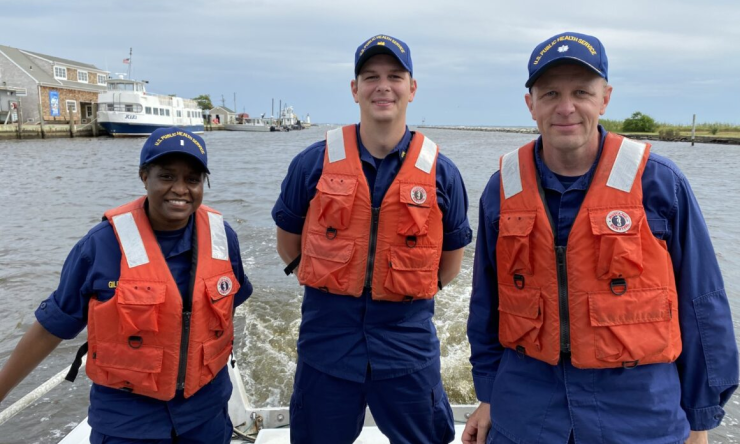 Three public health service officers dressed in dark blue uniforms with dark blue hats emblazoned with "U.S. Public Health Service", wearing bright orange life jackets on a boat in a harbor.